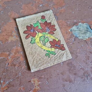 Puede incluir: Un parche rectangular con un dise&ntilde;o floral. El parche presenta flores rojas, hojas verdes y una herradura amarilla. El parche est&aacute; sobre un fondo marr&oacute;n con costuras blancas en los bordes. El fondo es una superficie desgastada, descascarada, de color marr&oacute;n rojizo.