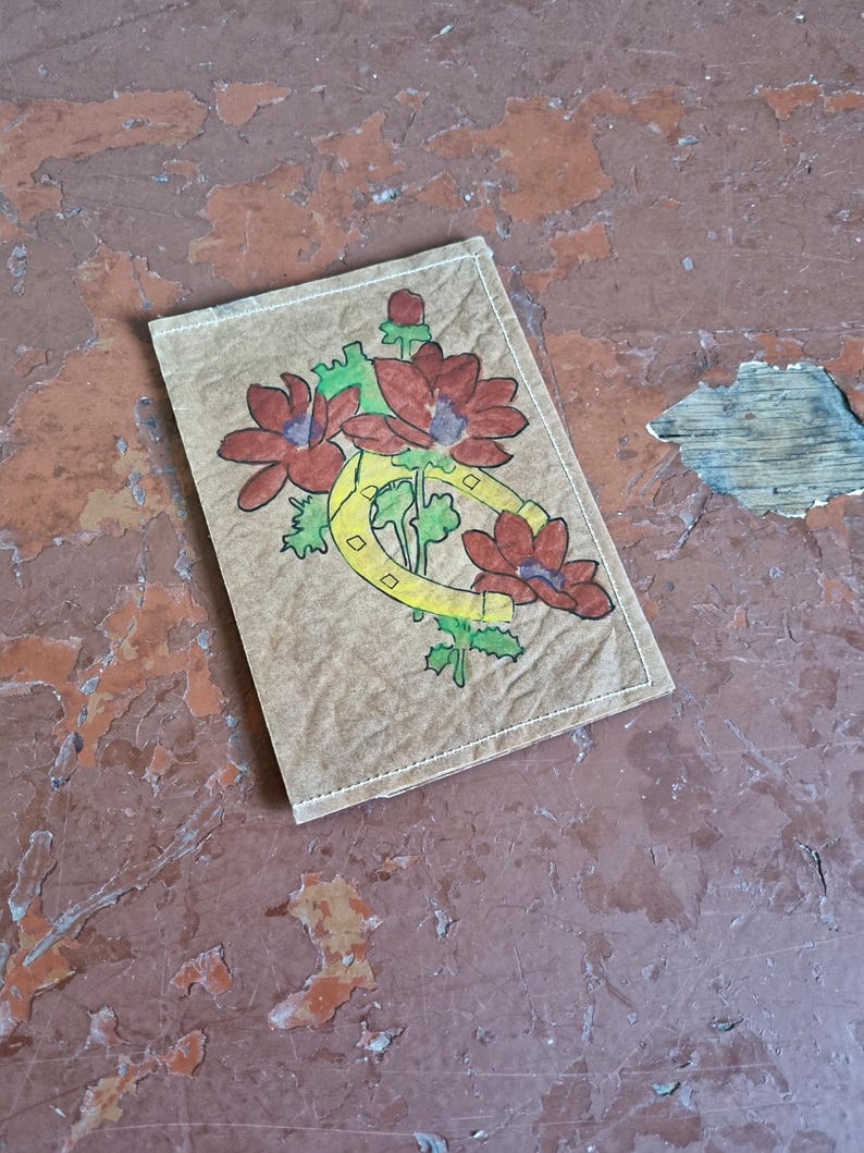 Puede incluir: Un parche rectangular con un dise&ntilde;o floral. El parche presenta flores rojas, hojas verdes y una herradura amarilla. El parche est&aacute; sobre un fondo marr&oacute;n con costuras blancas en los bordes. El fondo es una superficie desgastada, descascarada, de color marr&oacute;n rojizo.