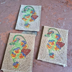 May include: Three rectangular, tan coasters with a floral design. Each coaster features a hand-painted image of a basket of pansies in yellow, orange, red, blue, and green. The coasters are made of a textured material and have white stitching around the edges.