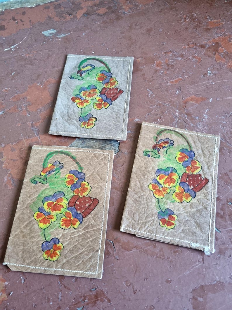 May include: Three rectangular, tan coasters with a floral design. Each coaster features a hand-painted image of a basket of pansies in yellow, orange, red, blue, and green. The coasters are made of a textured material and have white stitching around the edges.