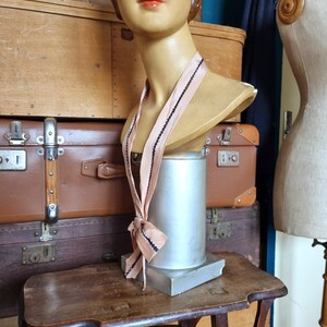 May include: A vintage mannequin head with a blush pink ribbon, accented with black trim, draped around its neck. The mannequin is displayed on a small wooden table, with a metal base. The background features stacked antique suitcases and a dress form.