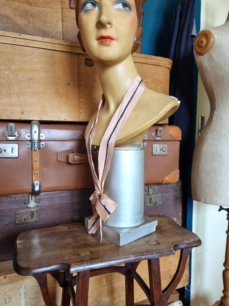 May include: A vintage mannequin head with a blush pink ribbon, accented with black trim, draped around its neck. The mannequin is displayed on a small wooden table, with a metal base. The background features stacked antique suitcases and a dress form.