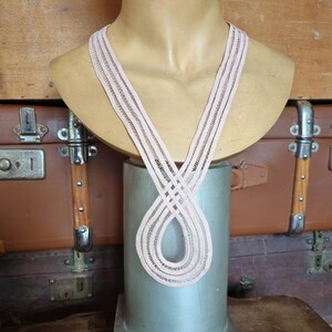 Peut inclure: Un collier rose clair en tissage ouvert pr&eacute;sent&eacute; sur un buste de mannequin. Le collier est fait d'un mat&eacute;riau tiss&eacute;, formant un motif en forme de huit. Le buste est pos&eacute; sur un pi&eacute;destal argent&eacute;, avec des bagages vintage en arri&egrave;re-plan.