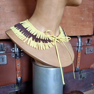 May include: A yellow and brown fringed collar necklace displayed on a mannequin bust. The collar is made of layered strips of yellow and brown material, tied with a yellow ribbon. The bust is on a silver stand.