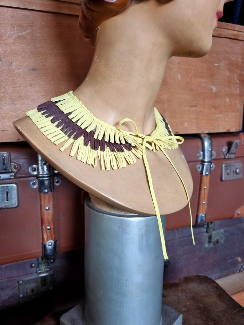 May include: A yellow and brown fringed collar necklace displayed on a mannequin bust. The collar is made of layered strips of yellow and brown material, tied with a yellow ribbon. The bust is on a silver stand.