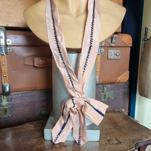 May include: A peach-colored necktie with dark blue stitching along the edges, tied in a bow. The tie is displayed on a mannequin bust. The background includes vintage luggage and a wooden table.