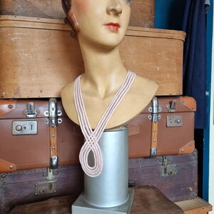 Peut inclure: Un buste de mannequin vintage avec des traits peints, portant un collier ray&eacute; rose clair et noir. Le mannequin est expos&eacute; sur un pi&eacute;destal argent&eacute;, avec des malles en bois antiques en arri&egrave;re-plan.