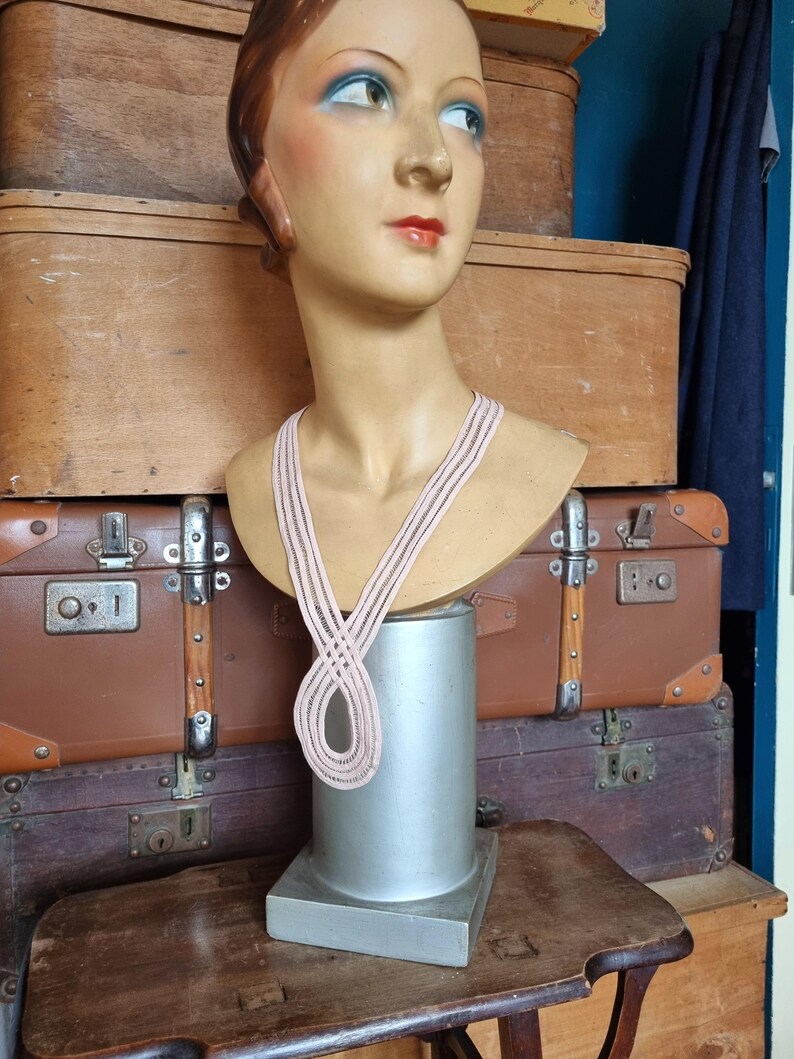 Peut inclure: Un buste de mannequin vintage avec des traits peints, portant un collier ray&eacute; rose clair et noir. Le mannequin est expos&eacute; sur un pi&eacute;destal argent&eacute;, avec des malles en bois antiques en arri&egrave;re-plan.
