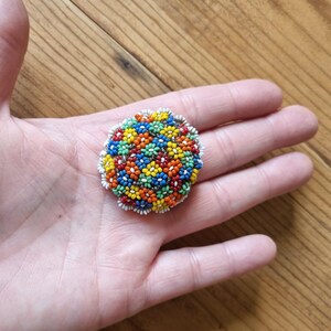 May include: A round beaded brooch with a floral design. The beads are in shades of red, orange, yellow, green, and blue. The brooch is surrounded by a white beaded border.