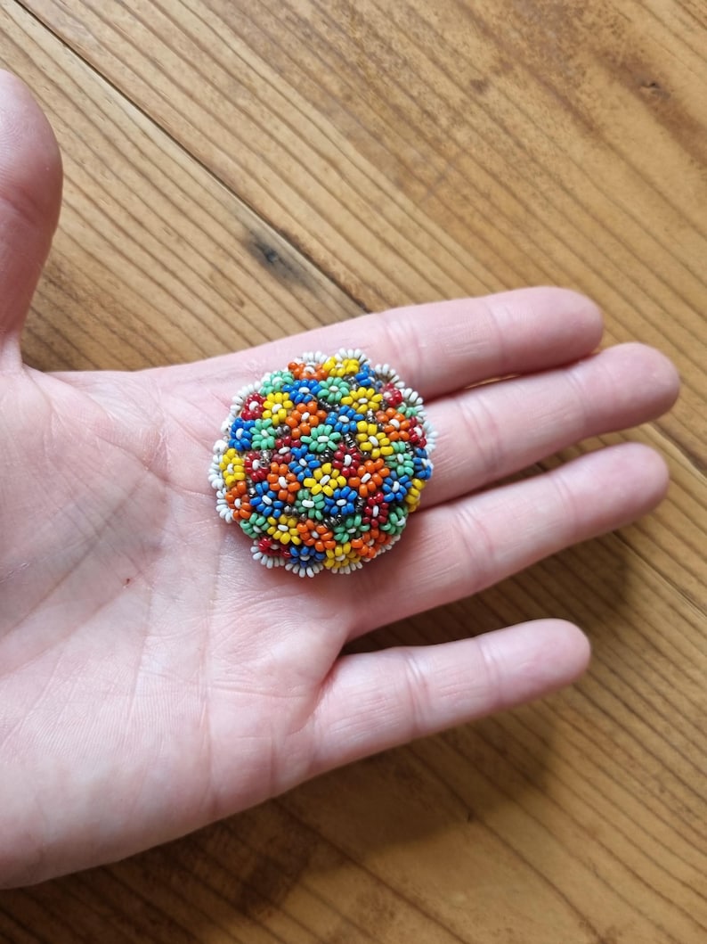 May include: A round beaded brooch with a floral design. The beads are in shades of red, orange, yellow, green, and blue. The brooch is surrounded by a white beaded border.