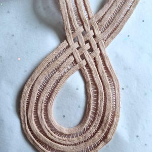 Peut inclure: Gros plan sur un ruban tiss&eacute; de couleur p&ecirc;che avec un motif en boucle. Le ruban est fait d'une mati&egrave;re douce et textur&eacute;e et pr&eacute;sente une s&eacute;rie de lignes parall&egrave;les et d'espaces ouverts. Le design est complexe et semble fait &agrave; la main.
