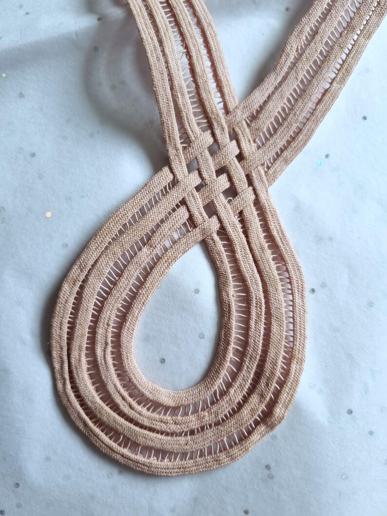 Peut inclure: Gros plan sur un ruban tiss&eacute; de couleur p&ecirc;che avec un motif en boucle. Le ruban est fait d'une mati&egrave;re douce et textur&eacute;e et pr&eacute;sente une s&eacute;rie de lignes parall&egrave;les et d'espaces ouverts. Le design est complexe et semble fait &agrave; la main.
