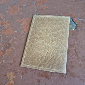 Puede incluir: Una cartera rectangular de cuero color marr&oacute;n claro con una superficie texturizada. Los bordes est&aacute;n cosidos con hilo blanco. La cartera est&aacute; sobre una superficie desgastada de color marr&oacute;n rojizo.