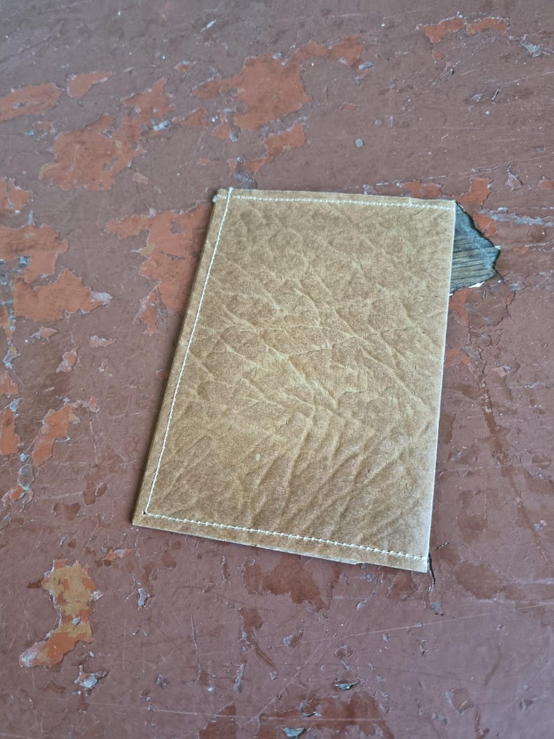 Puede incluir: Una cartera rectangular de cuero color marr&oacute;n claro con una superficie texturizada. Los bordes est&aacute;n cosidos con hilo blanco. La cartera est&aacute; sobre una superficie desgastada de color marr&oacute;n rojizo.