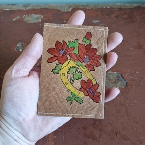 Puede incluir: Una peque&ntilde;a tarjeta rectangular marr&oacute;n con un dise&ntilde;o dibujado a mano con flores rojas, hojas verdes y una herradura amarilla. La tarjeta tiene un borde blanco cosido y se sostiene en una mano.
