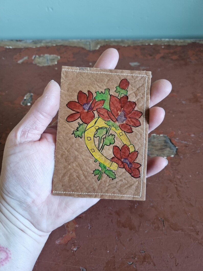 Puede incluir: Una peque&ntilde;a tarjeta rectangular marr&oacute;n con un dise&ntilde;o dibujado a mano con flores rojas, hojas verdes y una herradura amarilla. La tarjeta tiene un borde blanco cosido y se sostiene en una mano.