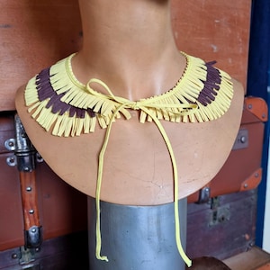 May include: A yellow and brown fringed collar necklace with a tie closure, displayed on a mannequin head. The collar features alternating sections of yellow and brown fringe, creating a decorative accessory.