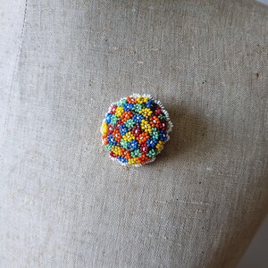 May include: A colorful beaded brooch in the shape of a circle. The brooch is made of small, round beads in shades of red, orange, yellow, green, blue, and white. The beads are arranged in a pattern of small flowers.