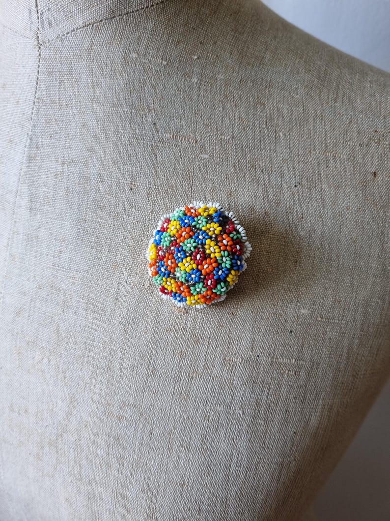 May include: A colorful beaded brooch in the shape of a circle. The brooch is made of small, round beads in shades of red, orange, yellow, green, blue, and white. The beads are arranged in a pattern of small flowers.