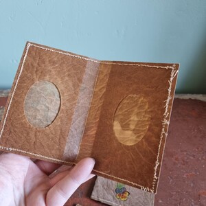 May include: A handmade brown paper photo album with two oval apertures for photos. The album is held open, revealing the interior and a clear strip down the centre. The edges are stitched with white thread. Small, decorated paper tags with floral designs are visible.