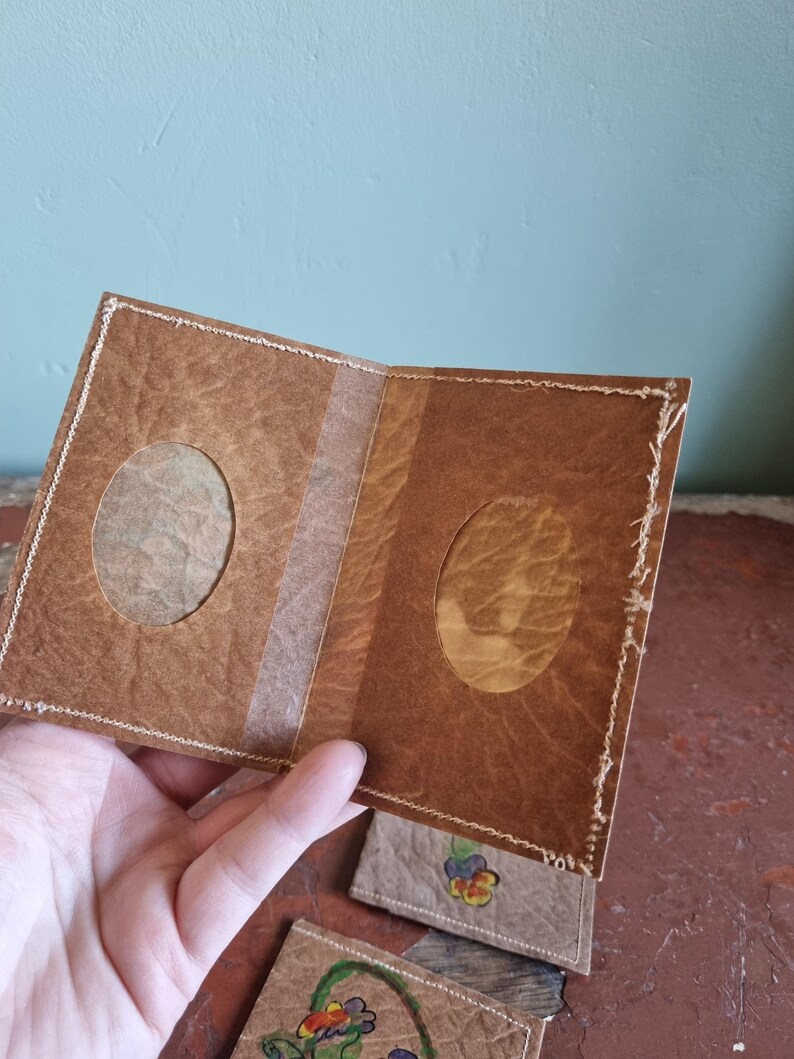 May include: A handmade brown paper photo album with two oval apertures for photos. The album is held open, revealing the interior and a clear strip down the centre. The edges are stitched with white thread. Small, decorated paper tags with floral designs are visible.