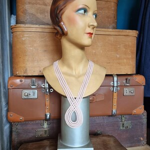Peut inclure: Un buste de mannequin vintage avec des traits peints, portant un collier rose clair. Le mannequin est expos&eacute; sur un socle argent&eacute;, avec en toile de fond des valises et des bo&icirc;tes en bois antiques. Le mannequin a les cheveux bruns et porte un collier rose clair.