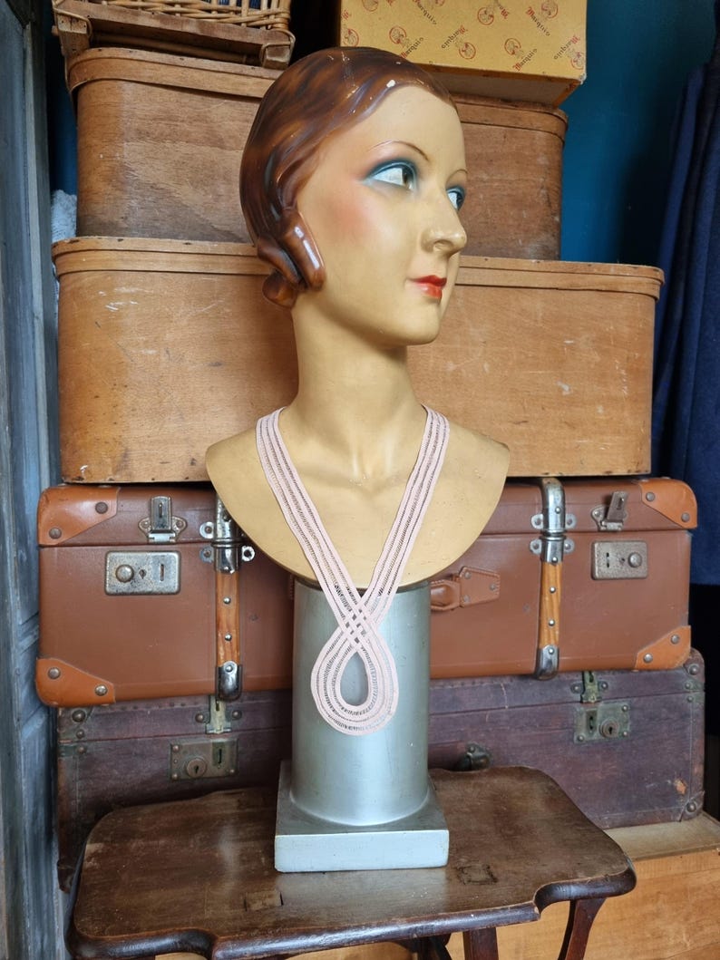 Peut inclure: Un buste de mannequin vintage avec des traits peints, portant un collier rose clair. Le mannequin est expos&eacute; sur un socle argent&eacute;, avec en toile de fond des valises et des bo&icirc;tes en bois antiques. Le mannequin a les cheveux bruns et porte un collier rose clair.