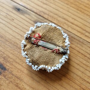 May include: A round, beige fabric pin with a white bead border. The pin has a metal clasp on the back.