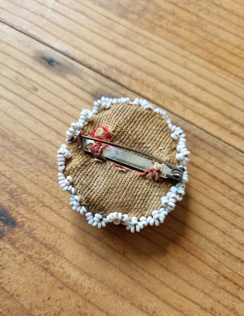 May include: A round, beige fabric pin with a white bead border. The pin has a metal clasp on the back.