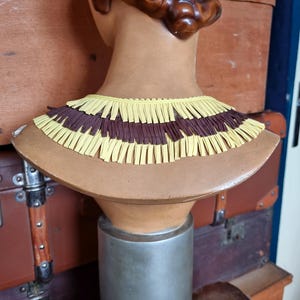 May include: A decorative bust with a brown wig and a tan neck. The bust is adorned with a layered collar featuring alternating rows of yellow and brown fringe. The bust is mounted on a silver stand.