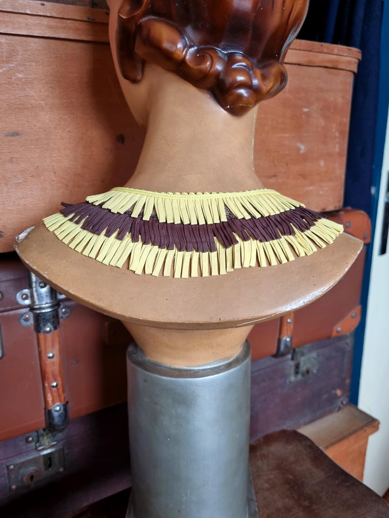 May include: A decorative bust with a brown wig and a tan neck. The bust is adorned with a layered collar featuring alternating rows of yellow and brown fringe. The bust is mounted on a silver stand.