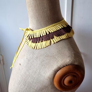 May include: A yellow and brown fringed collar displayed on a mannequin. The collar has a yellow base with alternating rows of yellow and brown fringe. A yellow tie secures the collar. The mannequin is beige with a wooden neck and shoulder.