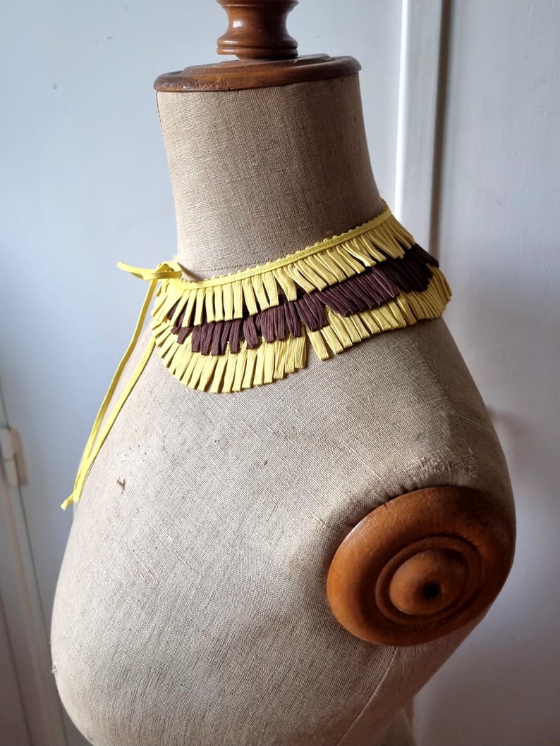 May include: A yellow and brown fringed collar displayed on a mannequin. The collar has a yellow base with alternating rows of yellow and brown fringe. A yellow tie secures the collar. The mannequin is beige with a wooden neck and shoulder.