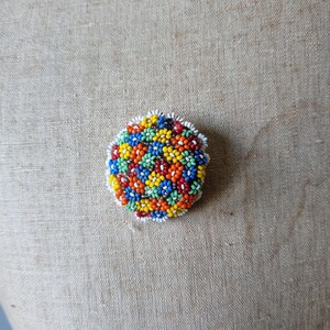 May include: A round, colorful beaded brooch with a floral pattern. The beads are in shades of red, orange, yellow, green, blue, and white.
