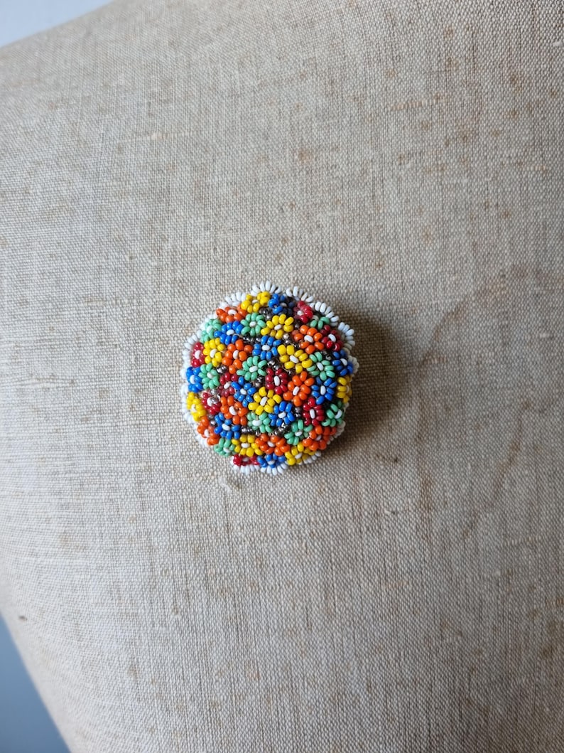 May include: A round, colorful beaded brooch with a floral pattern. The beads are in shades of red, orange, yellow, green, blue, and white.