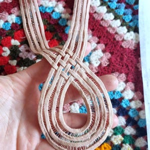 Peut inclure: Une garniture en dentelle beige tiss&eacute;e avec un motif en forme de goutte d'eau. La garniture est tenue dans une main, mettant en valeur le motif de tissage complexe. L'arri&egrave;re-plan est une couverture crochet&eacute;e color&eacute;e.