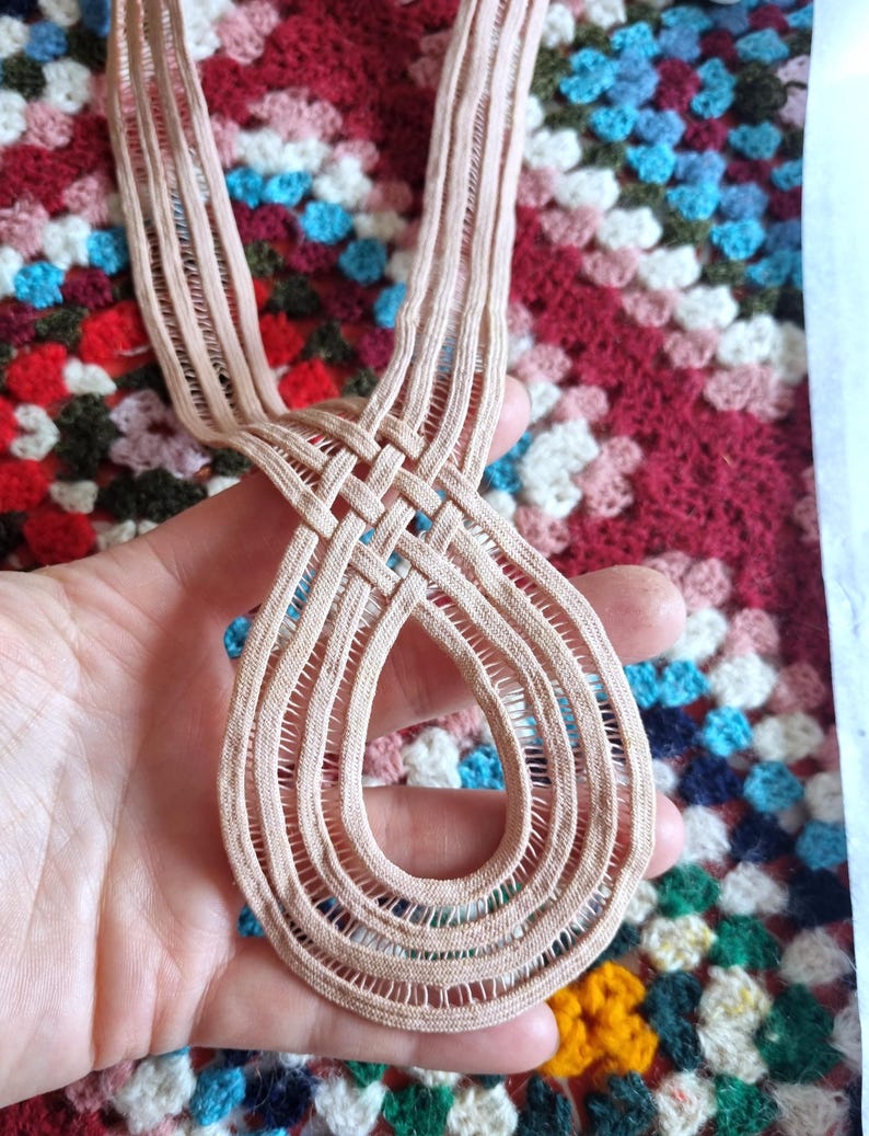 Peut inclure: Une garniture en dentelle beige tiss&eacute;e avec un motif en forme de goutte d'eau. La garniture est tenue dans une main, mettant en valeur le motif de tissage complexe. L'arri&egrave;re-plan est une couverture crochet&eacute;e color&eacute;e.