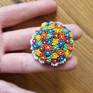 May include: A colorful beaded brooch with a floral design. The brooch is made of small, round beads in various colors, including red, orange, yellow, green, blue, and white. The beads are arranged in a circular pattern to create a flower-like design.