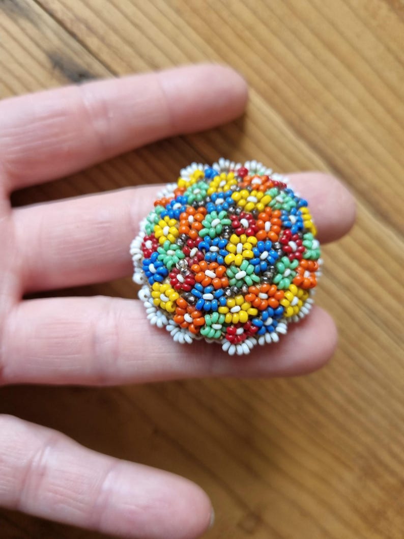 May include: A colorful beaded brooch with a floral design. The brooch is made of small, round beads in various colors, including red, orange, yellow, green, blue, and white. The beads are arranged in a circular pattern to create a flower-like design.