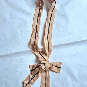 May include: A peach-colored satin ribbon with black stitching along the edges and a bow at the bottom. The ribbon is folded and tied into a bow shape, with the ends of the ribbon extending downwards. The stitching is visible and adds a decorative element.