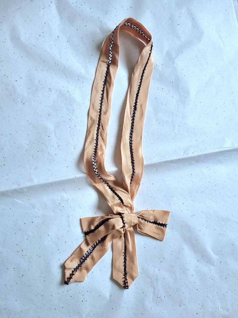 May include: A peach-colored satin ribbon with black stitching along the edges and a bow at the bottom. The ribbon is folded and tied into a bow shape, with the ends of the ribbon extending downwards. The stitching is visible and adds a decorative element.