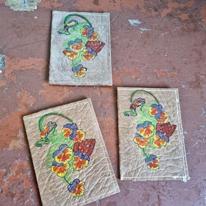 May include: Three rectangular coasters with a floral design. Each coaster has a beige background with a hand-painted image of pansies in yellow, orange, blue, and red, with green stems and leaves. The coasters have white stitching.