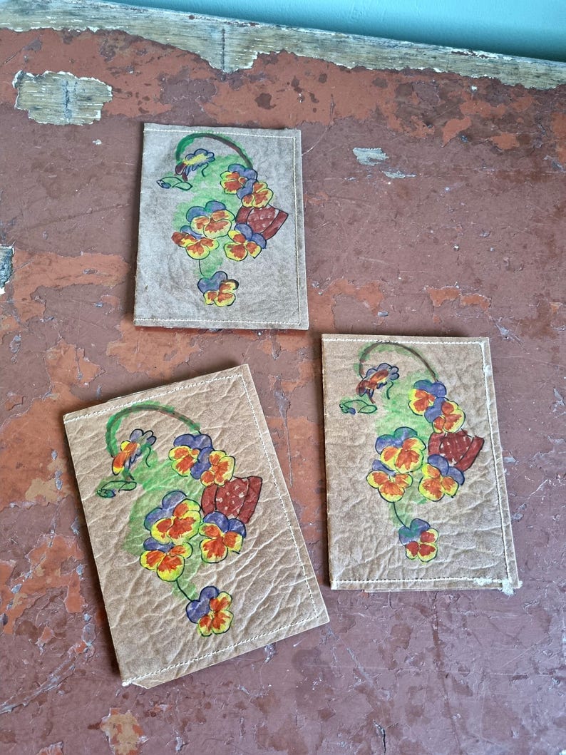 May include: Three rectangular coasters with a floral design. Each coaster has a beige background with a hand-painted image of pansies in yellow, orange, blue, and red, with green stems and leaves. The coasters have white stitching.
