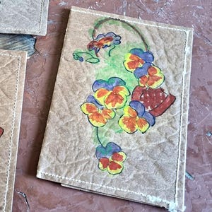 May include: A brown paper card with hand-painted floral art. The artwork depicts yellow, orange, and purple pansies with green stems. The card has a stitched border and is on a textured surface.