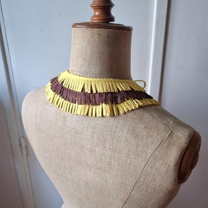 May include: A vintage-style collar made of yellow and brown fringed paper, displayed on a mannequin. The collar has a layered design with alternating bands of yellow and brown fringe. The mannequin is a neutral beige color.
