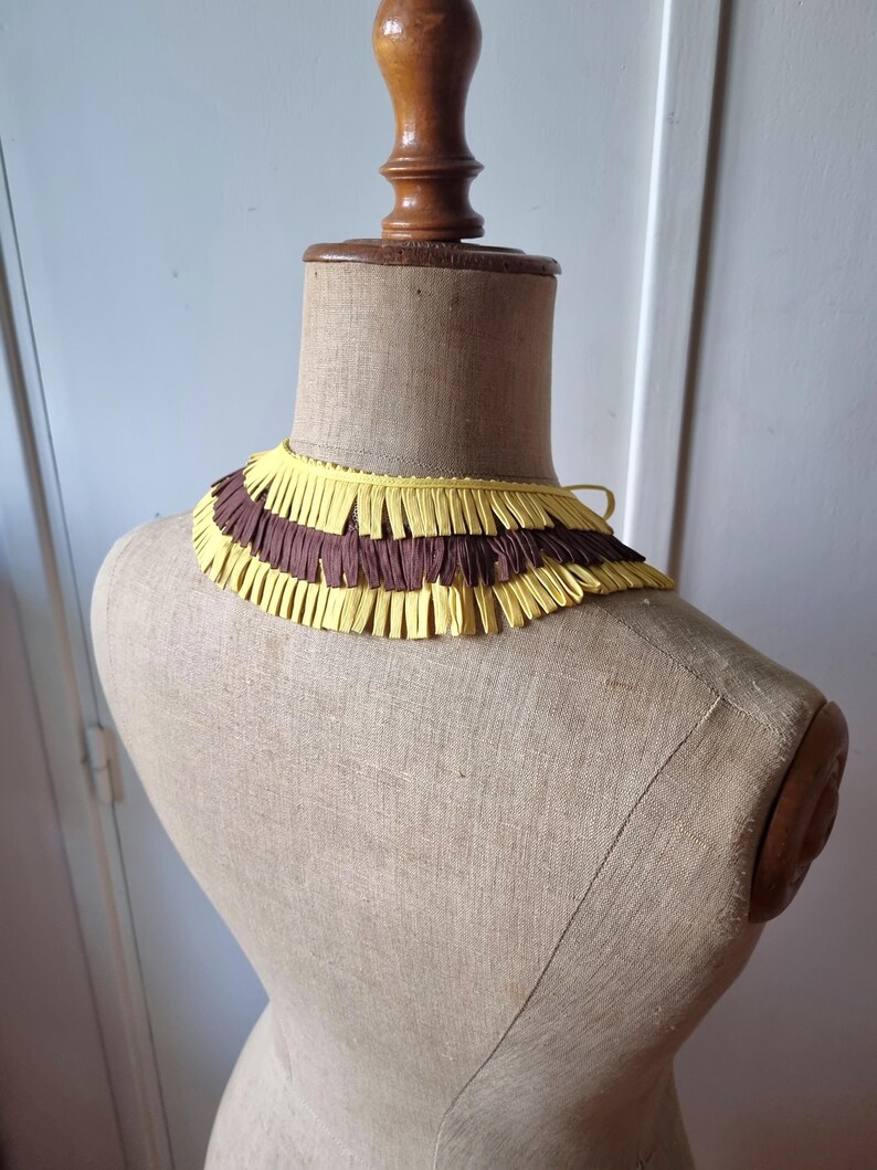 May include: A vintage-style collar made of yellow and brown fringed paper, displayed on a mannequin. The collar has a layered design with alternating bands of yellow and brown fringe. The mannequin is a neutral beige color.