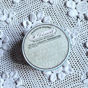 Puede incluir: Una caja redonda de hojalata blanca y azul con el texto "Pharmacie Dessort H. Dessort Pharmacien de 1ere Classe 35. Rue Lafayette, TOULOUSE TEL. 237-90" impreso en ella. La caja está sobre una superficie de crochet blanco.