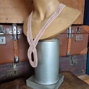 Peut inclure: Un collier tiss&eacute; rose clair est pr&eacute;sent&eacute; sur un buste de mannequin. Le buste est pos&eacute; sur un pi&eacute;destal argent&eacute;. L'arri&egrave;re-plan comprend des valises marron vintage et une table en bois.