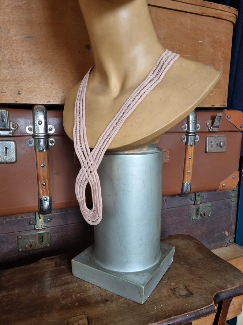 Peut inclure: Un collier tiss&eacute; rose clair est pr&eacute;sent&eacute; sur un buste de mannequin. Le buste est pos&eacute; sur un pi&eacute;destal argent&eacute;. L'arri&egrave;re-plan comprend des valises marron vintage et une table en bois.