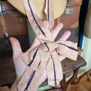 May include: A peach-colored satin ribbon with dark blue stitching detail. The ribbon is tied into a bow. The ribbon is likely vintage and is displayed against a vintage background.